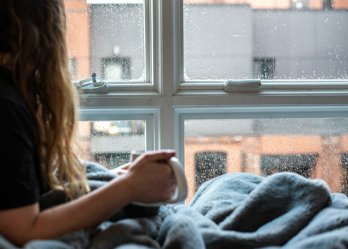 Person with a mug gazes out a rain-streaked window.